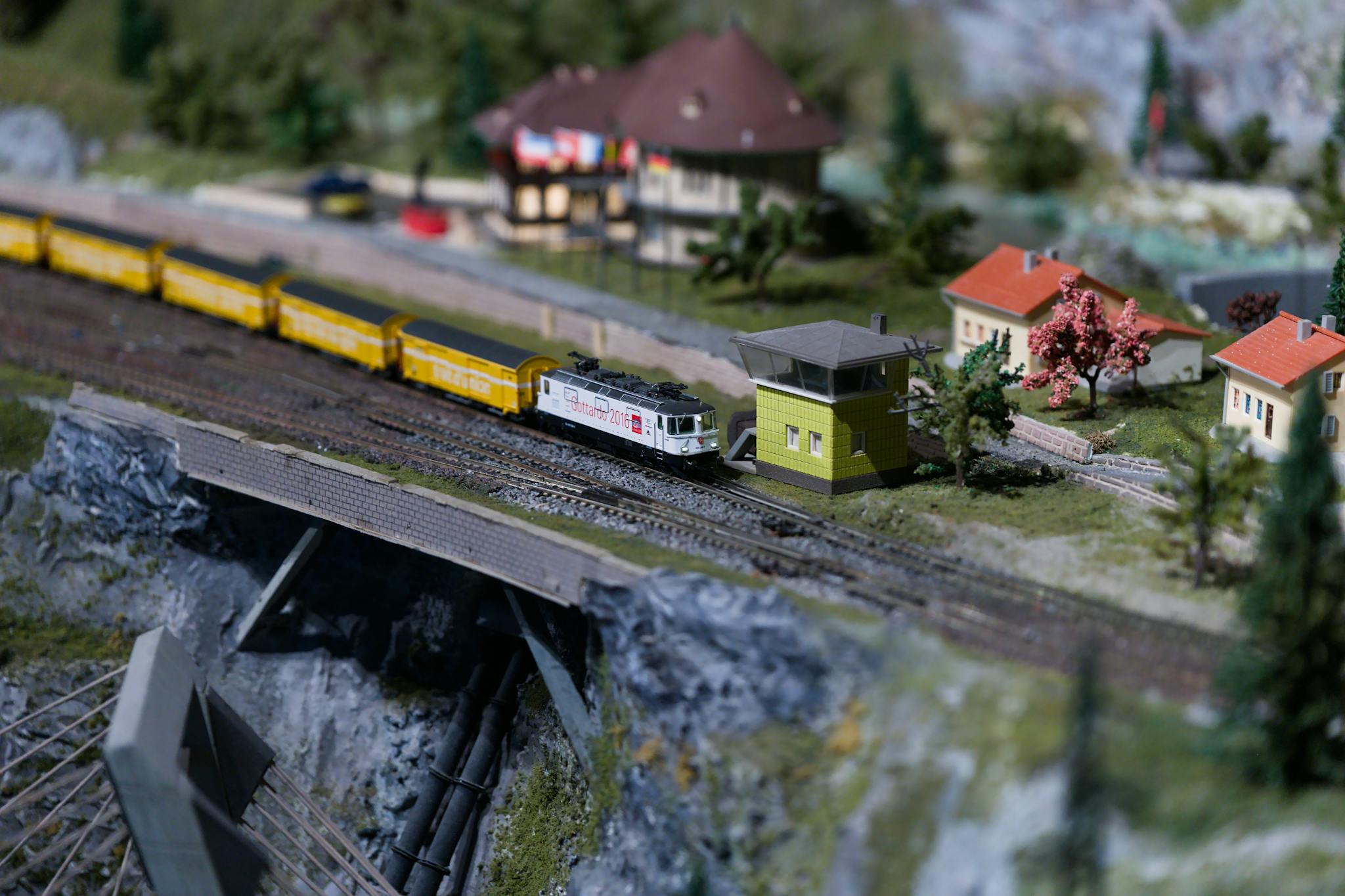 Detailed miniature model railway with yellow train crossing a bridge and small houses in the background.